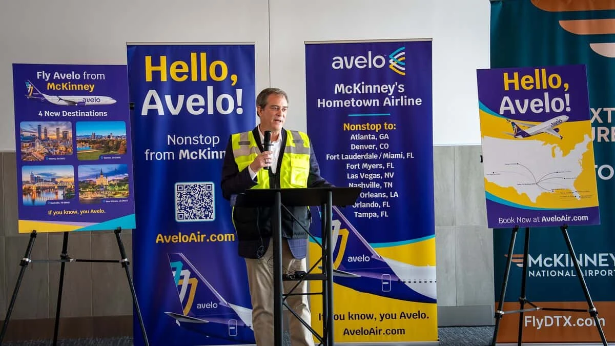 A speaker in a high-visibility vest at a lectern with a microphone, in front of Avelo Airlines banners and a McKinney National Airport banner.