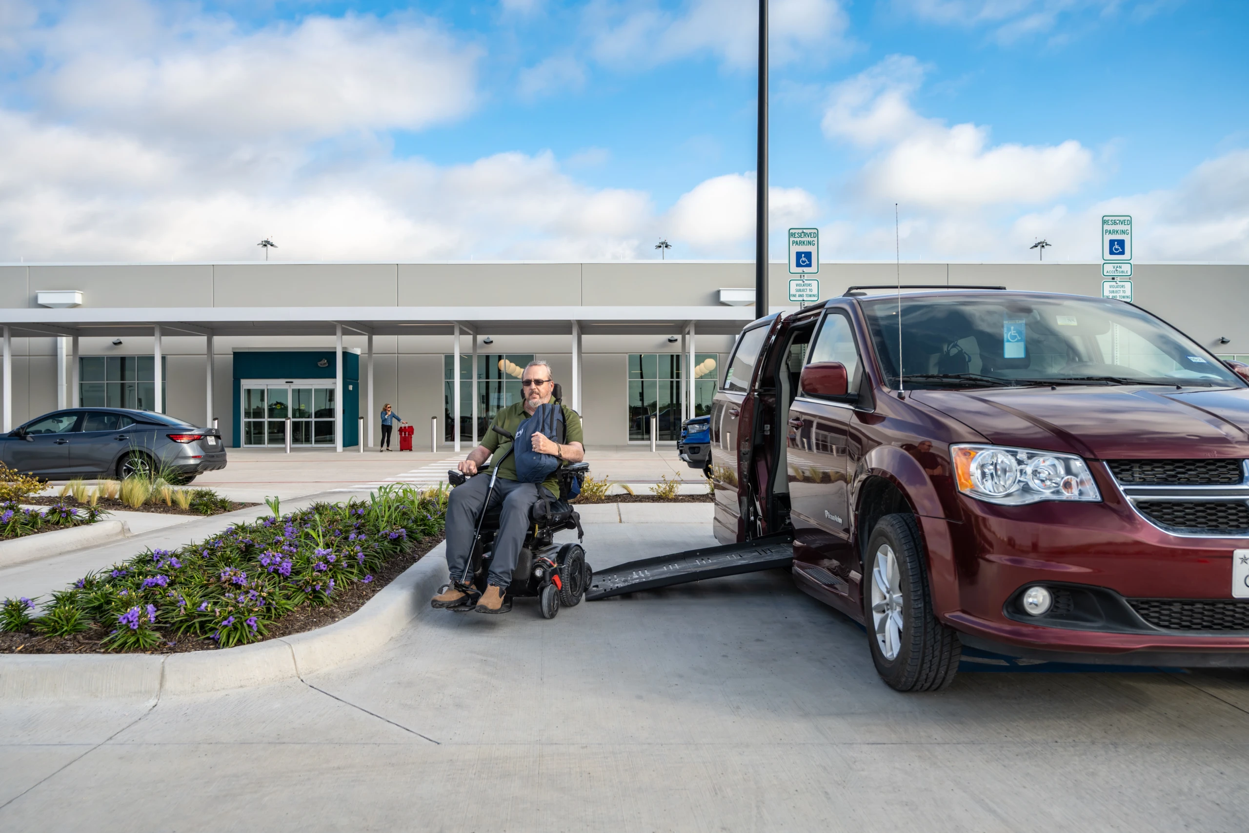 A traveler in a power wheelchair beside an accessible van with its ramp lowered, outside the McKinney National Airport terminal.