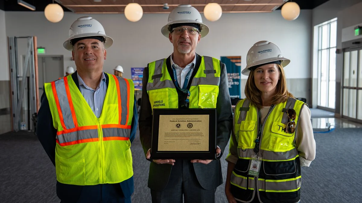 Three people in hard hats and safety vests inside the new McKinney National Airport passenger terminal; the man in the centre holds the framed FAA Airport Operating Certificate.