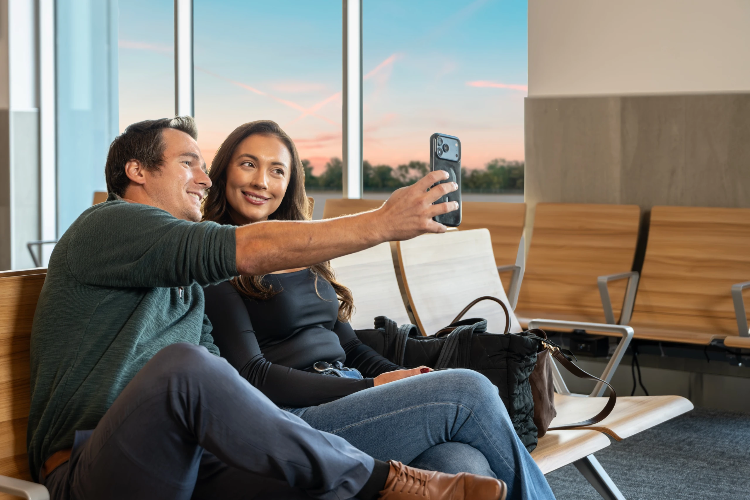 A couple takes a selfie in the gate seating as the sun sets outside the terminal windows.