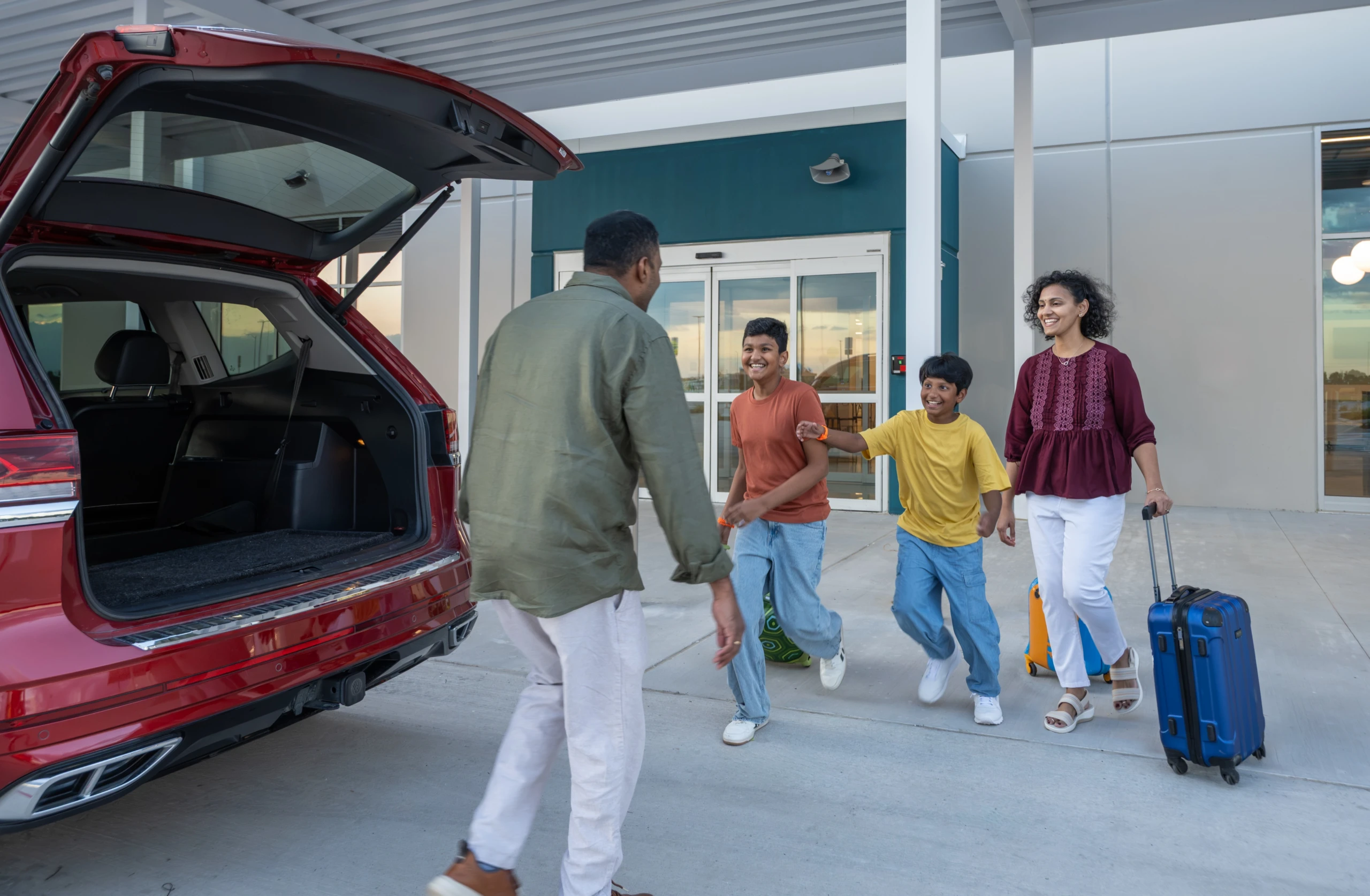 A family with suitcases greets a driver at the open trunk of an SUV outside the terminal doors.