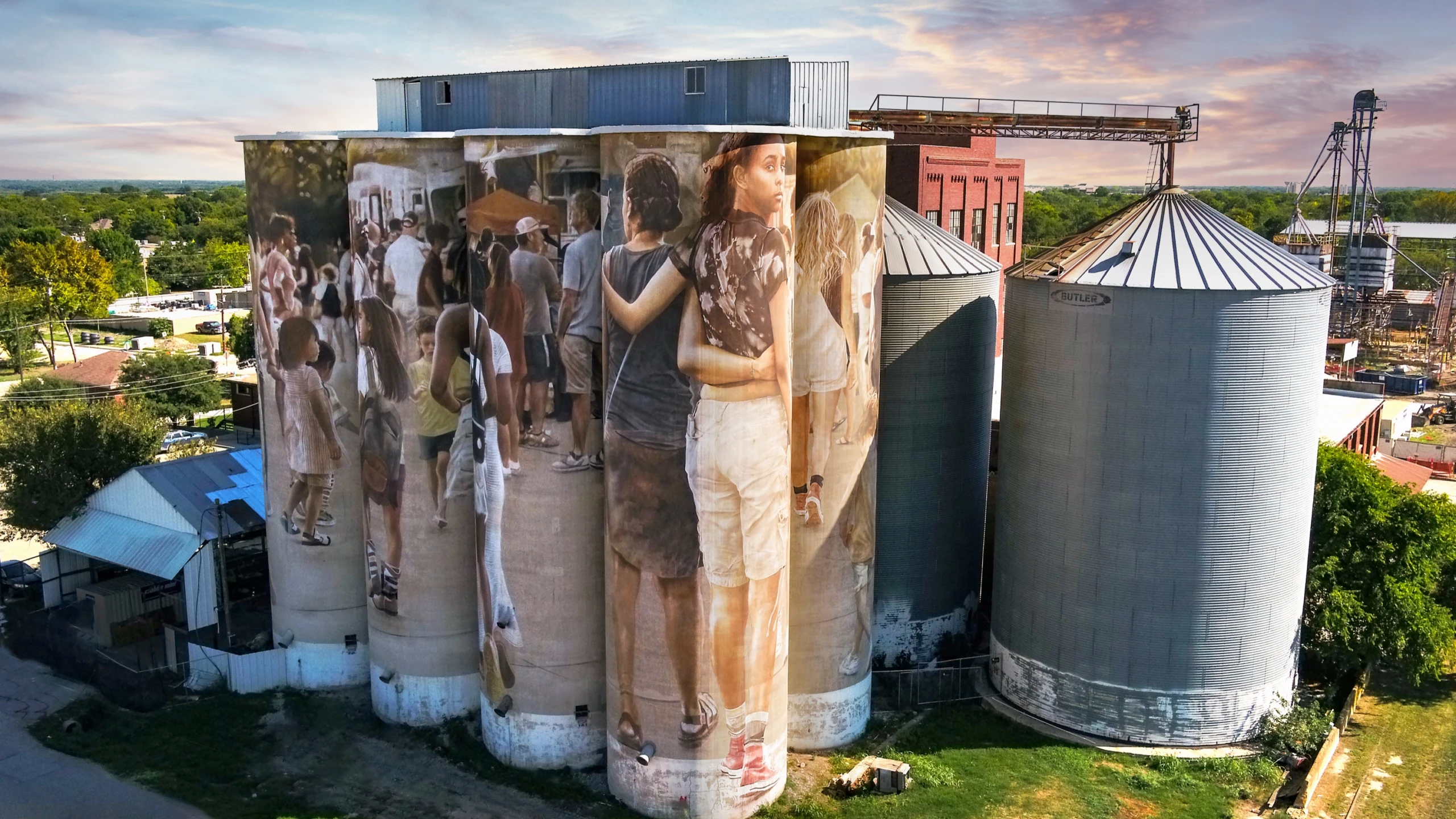 Grain silos in East McKinney painted with a large mural of people, seen from the air at sunset.